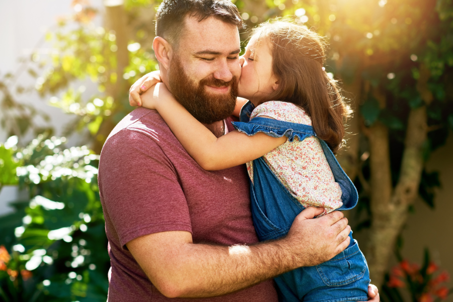 Un père portant sa fille qui lui fait un bisou dans le jardin. Un moment de tendresse entre un père et sa fille pour la Fête des Pères.