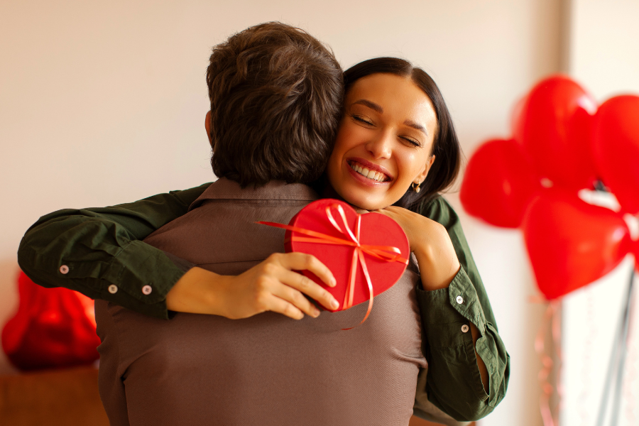 Femme souriante enlacant son partenaire lors de la Saint-Valentin, tenant un cadeau en forme de c&oelig;ur